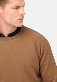 Rudas megztinis su apvaliu kaklu, raglano rankovėmis ir raukšlėtomis kraštinėmis, uždėtas ant juodos marškinėlių su apykakle. Matoma tekstūruota audinio detalė.