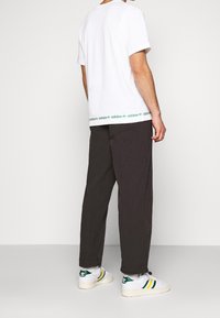 Vit bomullströja med grön Adidas-logga vid nederkanten, åtföljd av löst sittande mörkgrå byxor och vita sneakers med gröna och gula ränder.