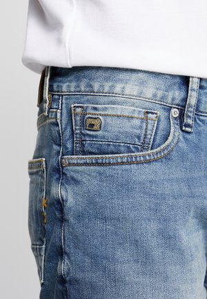 Gros plan sur la zone de la poche d'un jean denim bleu clair avec des coutures visibles, des rivets et une étiquette métallique avec logo, porté avec une chemise blanche.