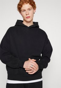 Sudadera negra con un bolsillo frontal, ajuste relajado, capucha ajustable con cordón, llevada sobre una camiseta blanca, con costuras visibles y puños acanalados.