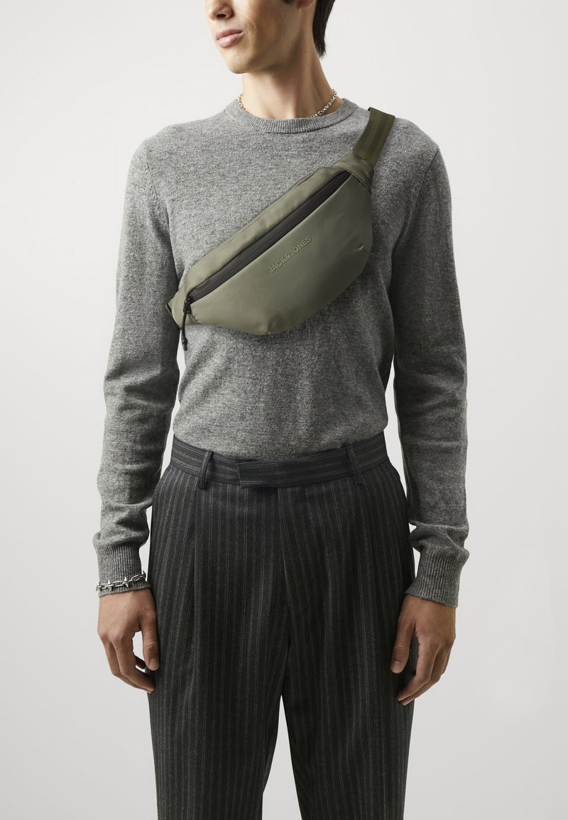 Bolsa de nylon verde en forma curva con un bolsillo con cremallera, llevada en bandolera. Presenta un logo de marca y una correa ajustable.