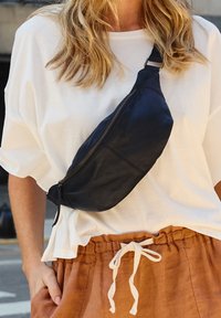 Marsupio in pelle nera con una forma curvata e una chiusura con zip, indossato di traverso sul petto di una persona, abbinato a una camicia bianca e pantaloni corti marroni.