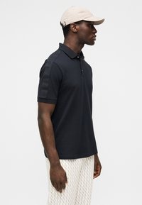 Granatowa koszulka polo z klasycznym kołnierzykiem, dwukolorowym zapięciem na guziki i fakturowaną tkaniną. Ozdobiona ciemnymi paskami po bokach. Jasnobeżowa czapka.