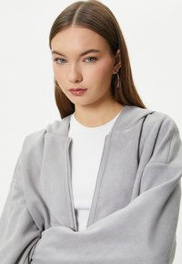 Grå lynlås-hættetrøje lavet af blødt materiale, med hætte og afslappet pasform, båret over en hvid ribbet skjorte. Glat tekstur og minimalistisk design.
