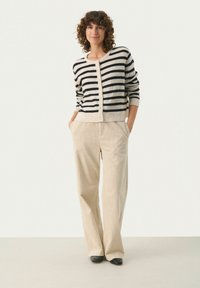 Zwart en crème gestreepte cardigan met knoopsluiting, gecombineerd met beige wijde corduroy broek. Model staat met handen in de zakken.