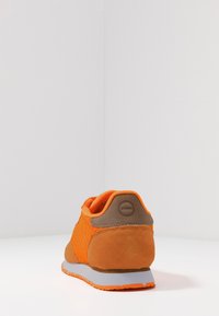 Orange idrottssko som visar mesh- och mockateksturer, med en grå sula och varumärke på hälen, placerad mot en enfärgad bakgrund.