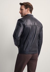 Czarna skórzana kurtka o gładkiej fakturze, z wyraźnie zaznaczonymi szwami i prostym kołnierzem. Założona na ciemnobrązowe spodnie, widok od tyłu.