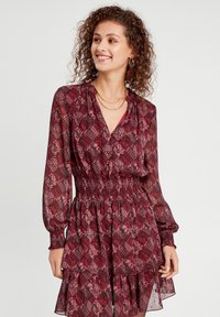 Robe à manches longues, décolleté en V, en bordeaux avec des motifs floraux et géométriques, taille élastiquée et volants superposés au niveau de l'ourlet. Texture en tissu transparent.