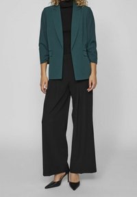 Blazer sarcelle, coupe décontractée avec manches retroussées, associé à un pantalon noir à larges jambes et des talons pointus noirs. Haut texturé noir en dessous.