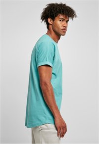 Camiseta de algodón turquesa con mangas enrolladas, cuello redondo clásico y corte relajado. La tela tiene una textura suave y una forma ligeramente holgada.