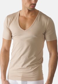 Man som bär en tight beige kortärmad V-ringad T-shirt och vita byxor, sedd från axlarna till midjan mot en vit bakgrund.
