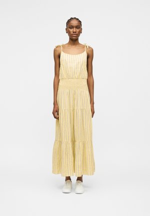 Ψηλή γυναίκα με πλεξούδες φοράει έναν κίτρινο μακρύ φορέμα χωρίς μανίκια με λεπτές μπλε κατακόρυφες ρίγες και λευκά αθλητικά παπούτσια, στέκεται μπροστά σε ένα μονόχρωμο φόντο.