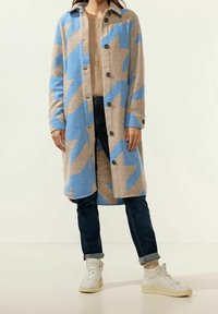 Lång kappa i beige med blått sicksackmönster, tillverkad av mjuk ull. Har en krage och knappstängning, tillsammans med mörka jeans och vita sneakers.