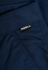 Navy blue denim fabric with a ribbed texture; features a black label with white text reading "REELL" near the waistband.