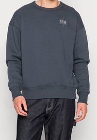 Mörkgrå sweatshirt med rund halsringning i en bomullsmix. Har lång ärm, ribbade muddar och nederkant, samt en liten logopatch på bröstet.