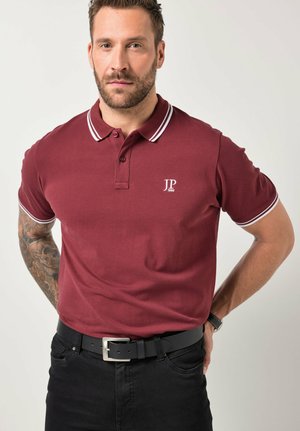 Mand med skæg og tatoveret venstre arm iklædt en bordeaux poloshirt med hvide kanter, sorte bukser og sort bælte, stående med hænderne bag ryggen.