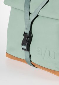 Borsa di stoffa verde con una base in pelle marrone. Presenta una fibbia in plastica nera e spallacci regolabili. Logo impresso sul lato.