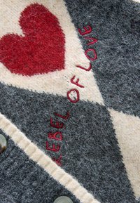 Grå och krämfärgad filt med en röd hjärtform och broderingen "REBEL OF LOVE", med en knappdetalj längs kanten. Rutinmönsterdesign.