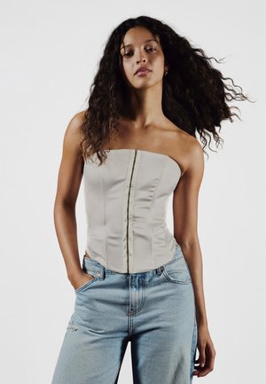 Woman with long curly hair wearing a strapless beige corset top and light blue jeans with her hands in pockets against a plain white background.