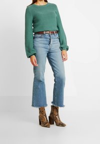 Mintgrön texturerad tröja med långa ärmar, kombinerad med ljusblå jeans med fransar. Brun läderbälte och bruna stövlar med ormskinnsmönster och klack.