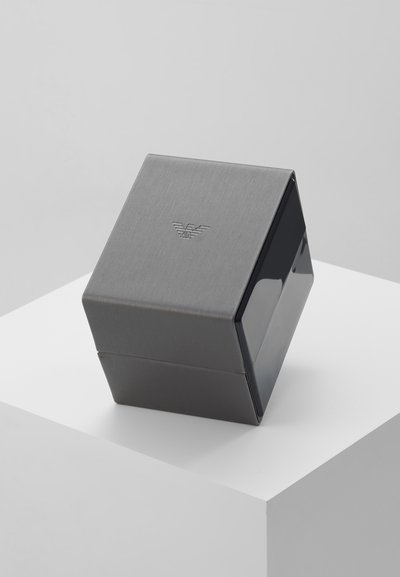 Caja cuadrada metálica gris con acabado cepillado y lados brillantes. Presenta un logotipo en relieve. Diseño compacto con bordes limpios y detalles mínimos.