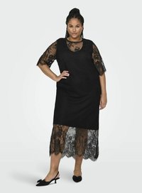 Vestito nero in pizzo con maniche corte, caratterizzato da un fondo in pizzo trasparente e orlo a scallop. Modello che indossa scarpe slingback nere con dettaglio a fiocco.