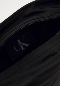 Interno della borsa in nylon nero con chiusura a zip. Presenta un logo "CK" in rilievo su un'etichetta in tessuto. Texture liscia, design minimalista.
