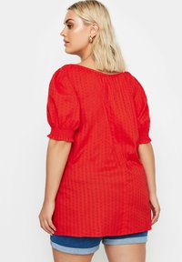 Rotes Texturoberteil mit Puffärmeln, elastischen Bündchen und lässigem Schnitt. Über Jeansshorts getragen, zeigt es einen runden Halsausschnitt und ein dezentes Muster.