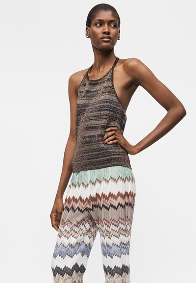 Γυναίκα που φοράει αμάνικο καφέ ριγέ τοπ halter και πολύχρωμο παντελόνι με φαρδιά πόδια και ζιγκ-ζαγκ σχέδιο, στέκεται με ένα χέρι στη μέση.