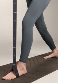 Sive leggins in joga trak za gležnjem, položen na goli nogi, ki počiva na strukturirani rjavi joga podlogi z merilnimi oznakami.