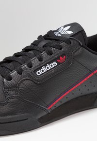 Primer plano de una zapatilla Adidas negra con cuero texturizado, cordones negros, rayas rojas y azules, y el logo de Adidas en el costado y en la lengua.