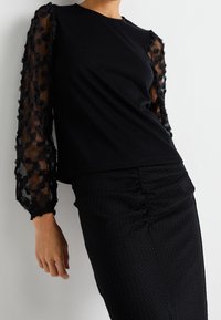 Svart topp med enfärgad kropp och genomskinliga svarta ärmar med blomstersbroderi, kombinerad med en svart kjol med strukturerade detaljer och rynkningar.