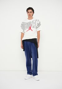 Fehér póló fekete és piros grafikai dizájnnal és logóval. Fekete réteges részlet a deréknál. Kék velúr nadrág és fehér tornacipő.