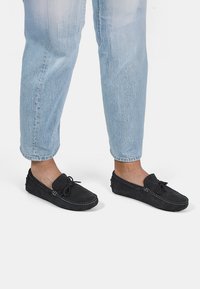 Melni zamšaini loafers ar izšūtu dizainu un loku detaļām, saskaņoti ar gaiši ziliem īsā kāja džinsiem, atklājot teksturētu audumu.