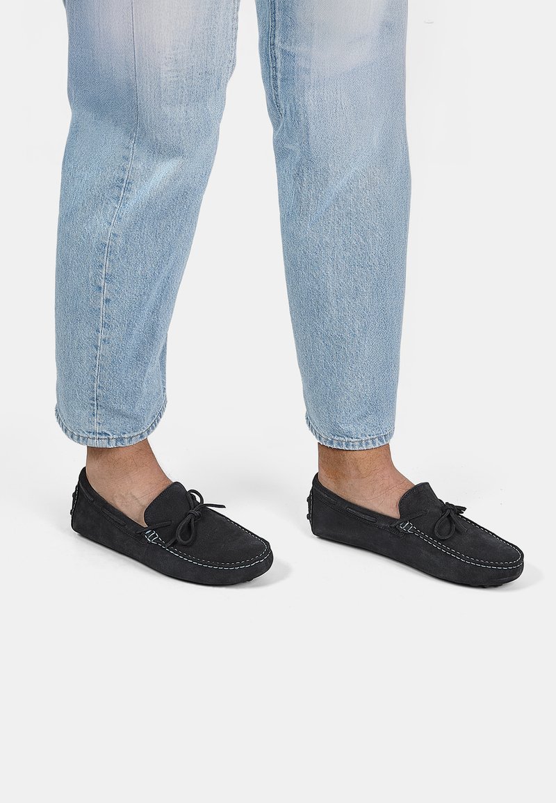 Melni zamšaini loafers ar izšūtu dizainu un loku detaļām, saskaņoti ar gaiši ziliem īsā kāja džinsiem, atklājot teksturētu audumu.