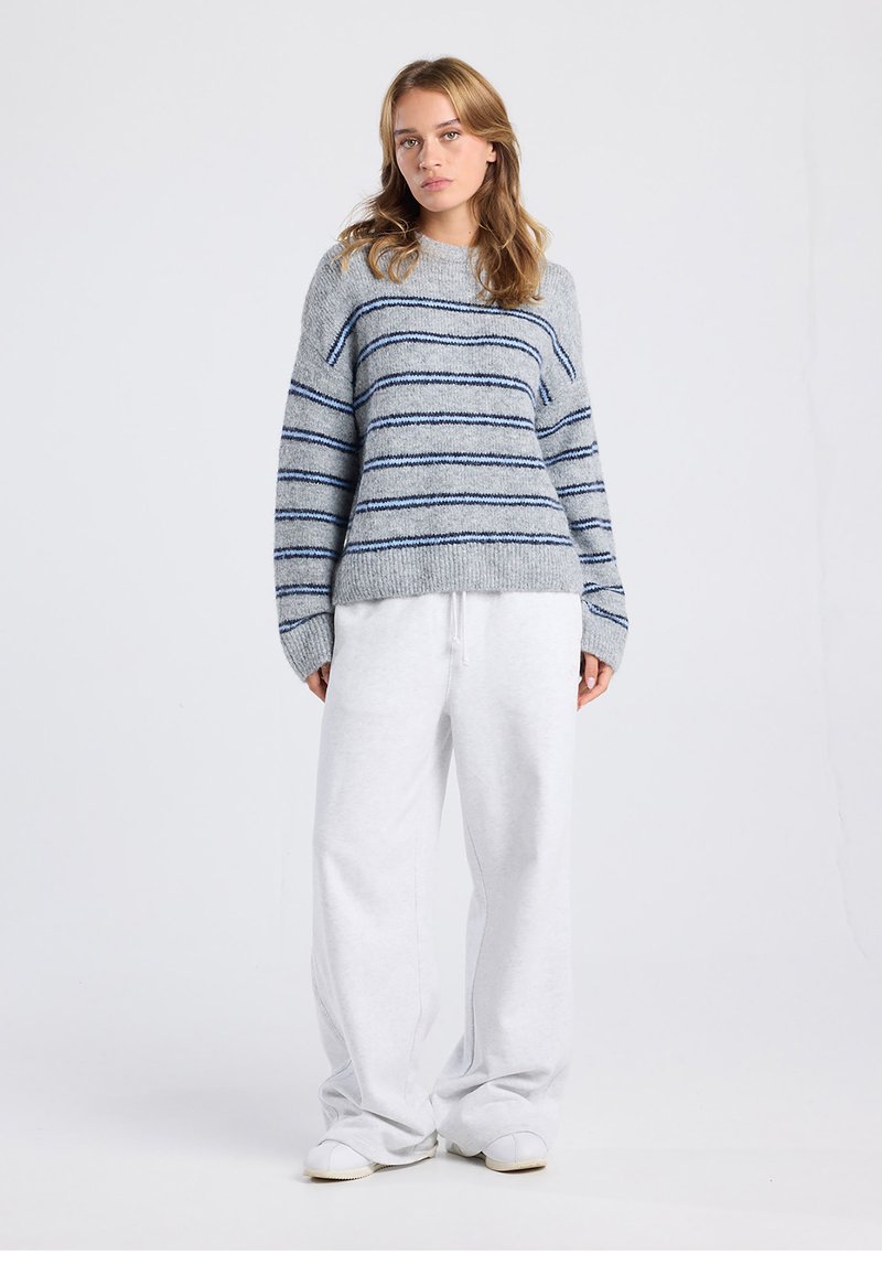 Hellgrauer Strickpullover mit marineblauen horizontalen Streifen, rundem Ausschnitt und überschnittenen Schultern, kombiniert mit locker sitzenden weißen Jogginghosen.