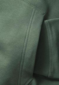 Gros plan sur un tissu vert montrant des coutures détaillées et un poignet côtelé sur une manche de vêtement pliée.