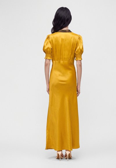 Femme aux longs cheveux foncés porte une robe en satin jaune moutarde avec des manches bouffantes et un col haut, associée à des talons hauts beiges, vue de dos.