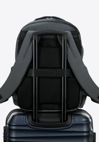 Sac à dos gris avec mesh noir et sangles, doté d'une poignée en métal rétractable, posé sur une valise rigide bleue. Logo sur la sangle.