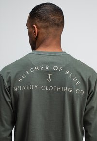 Man met kort haar, draagt een groen shirt met vervaagd witte tekst "Butcher of Blue Quality Clothing Co" op de achterkant, tegen een effen achtergrond.