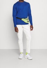 Grüner Hüftbeutel mit schwarzem Riemen, getragen über einem blauen Sweatshirt und weißen Hosen. Weiße Turnschuhe und leuchtend grüne Socken sind sichtbar.