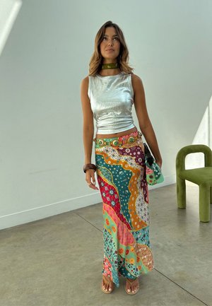 Γυναίκα με ασημί αμάνικη μπλούζα, πολύχρωμη φουσκωτή φούστα με σχέδια, πράσινο κολιέ και σανδάλια, στέκεται μέσα σε εσωτερικό χώρο κοντά σε πράσινη καρέκλα.