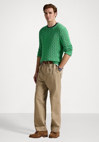 Pulover verde cu tricotaje, pantaloni bej și pantofi maro. Puloverul are guler rotund și mansete cu dungi. Ținută casual prezentată într-o poziție în picioare.