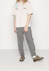 Beige bavlněné tričko s černým textem, šedé pruhované kalhoty a světle šedé boty s hnědými podrážkami. Model stojí s rukama podél těla.
