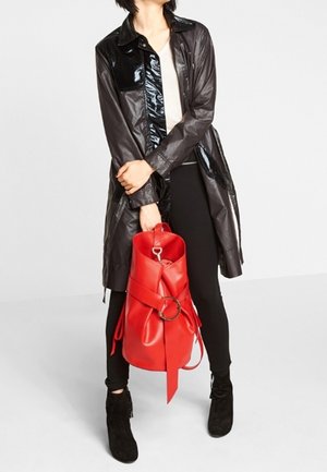 Trench-coat noir brillant, haut crème, pantalon noir et bottines noires. Tenant un sac à dos en cuir rouge avec un anneau circulaire et une sangle.
