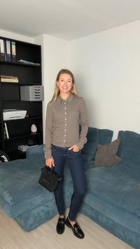 Grijs overhemd met knopen en kraag, donkerblauwe spijkerbroek en zwarte schoenen. Houdt een kleine zwarte handtas vast en staat in een kamer met een blauwe bank.