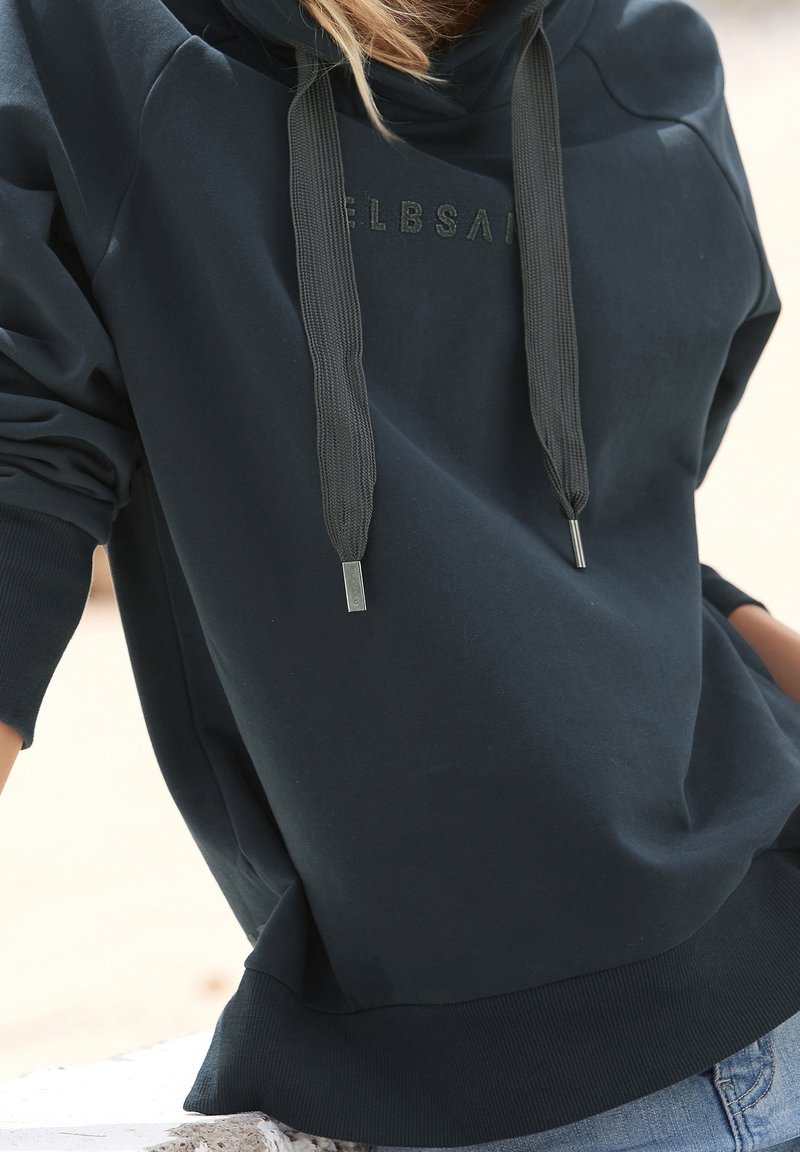 Personne portant un sweat à capuche oversize bleu marine foncé avec des cordons épais et un texte subtilement embossé, associé à un jean bleu clair.