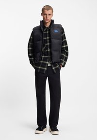 Μαύρο αμάνικο μπουφάν με ψηλό γιακά, φορεμένο πάνω από ένα σκοτεινό πράσινο καρό πουκάμισο, συνδυασμένο με μαύρο παντελόνι και μαύρα παπούτσια με κρέμ σόλες.