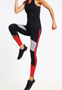 Černé tílko v kombinaci s úzkými legínami s černými, červenými a šedými panely. Černé sportovní boty dotvářejí celý outfit.
