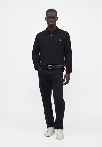 Homme debout portant un polo noir à manches longues, un pantalon noir avec ceinture et des baskets blanches sur fond blanc uni.